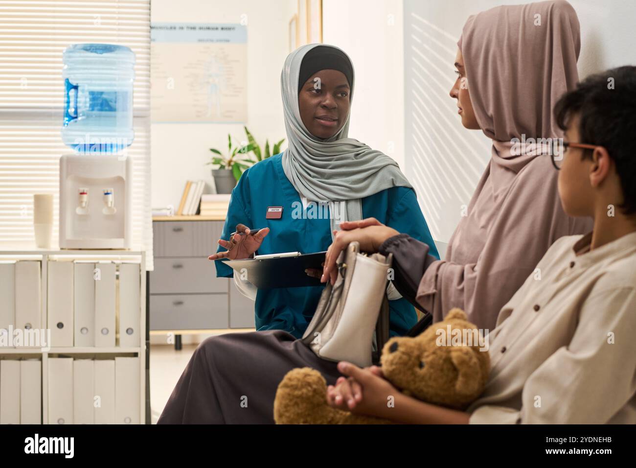 Professionnel de santé musulman en hijab consulte la famille dans le cabinet médical, engageant une conversation sur le bien-être du patient, assis à côté du distributeur d'eau Banque D'Images