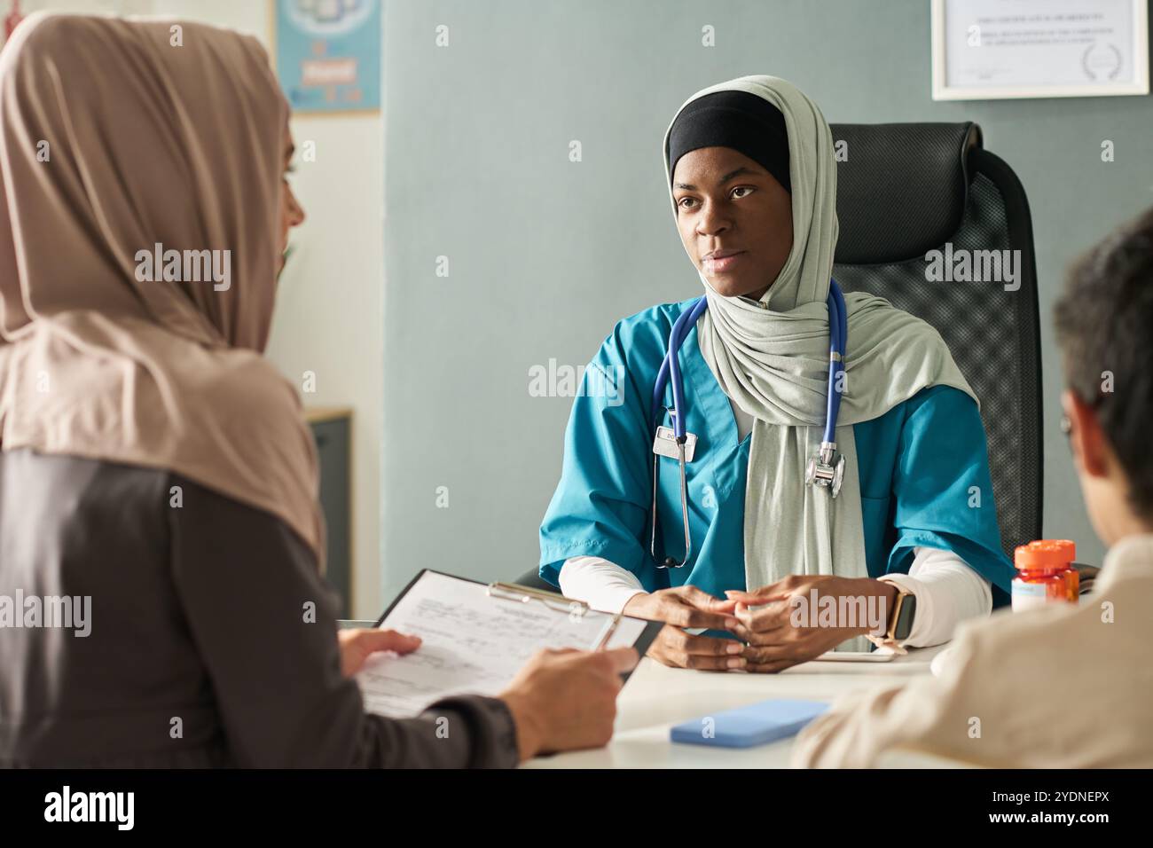 Fournisseur de soins de santé musulman s'engageant avec les patients, discutant du diagnostic médical, portant un stéthoscope et des gommages médicaux, assis dans un bureau clinique, axé sur les soins aux patients Banque D'Images