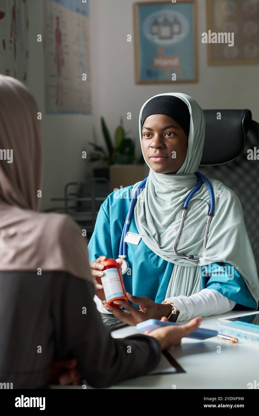 Portrait d'un professionnel de santé musulman en gommage consultant un patient en cabinet. Le médecin tient une bouteille de prescription tout en engageant la conversation Banque D'Images