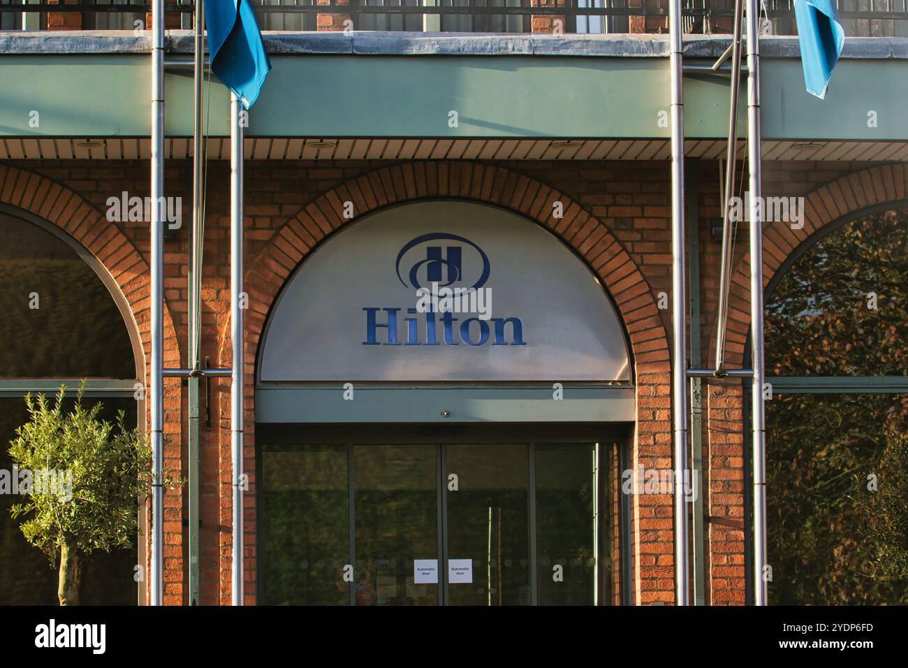 Un bâtiment en briques avec une grande entrée voûtée. L'entrée est surmontée d'une plaque argentée avec un logo Hilton bleu. Il y a deux drapeaux suspendus au-dessus Banque D'Images