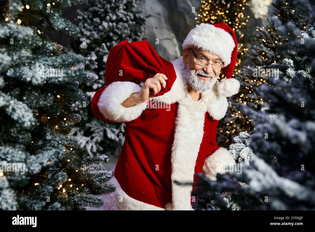 Un joyeux Père Noël avec un sac plein de cadeaux se promène parmi les arbres de Noël enneigés. Banque D'Images