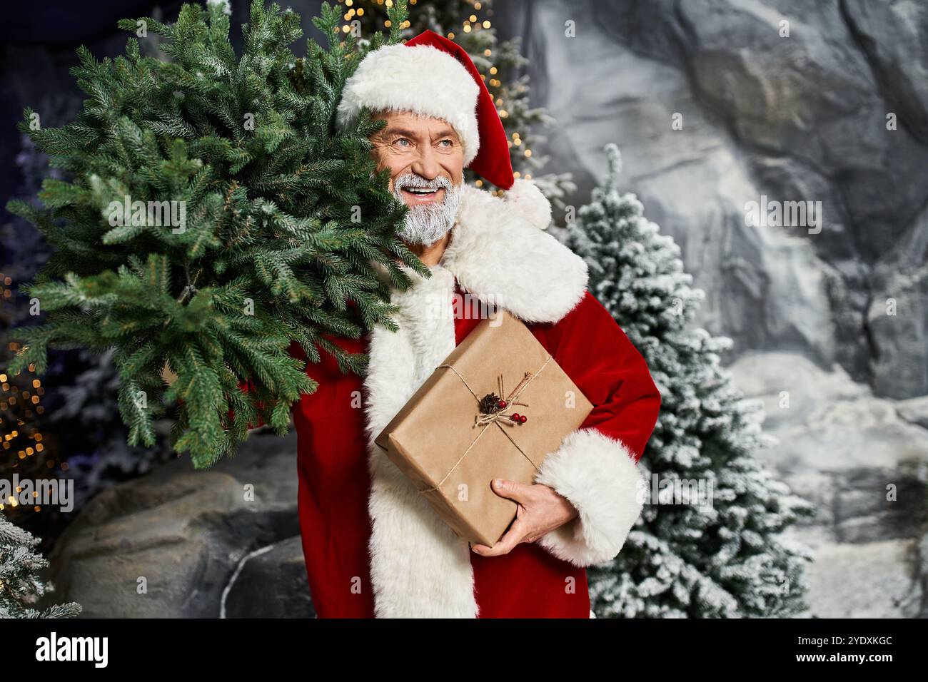 Le Père Noël tient un cadeau festif tout en étant entouré d'arbres couverts de neige et de décorations Banque D'Images