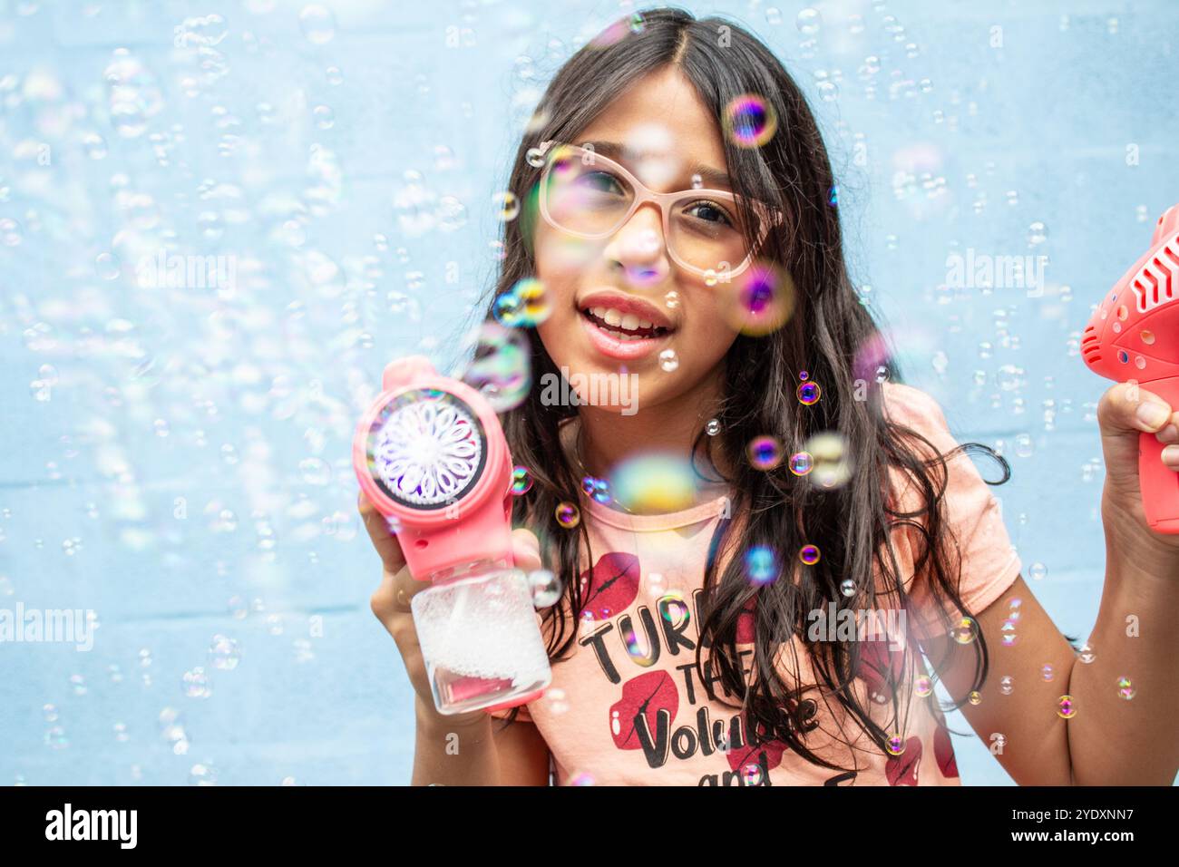 Goiania, Goias, Brésil – 13 octobre 2024 : gros plan du visage d'une jeune fille heureuse, jouant avec des bulles de savon, avec un mur bleu en arrière-plan. Banque D'Images