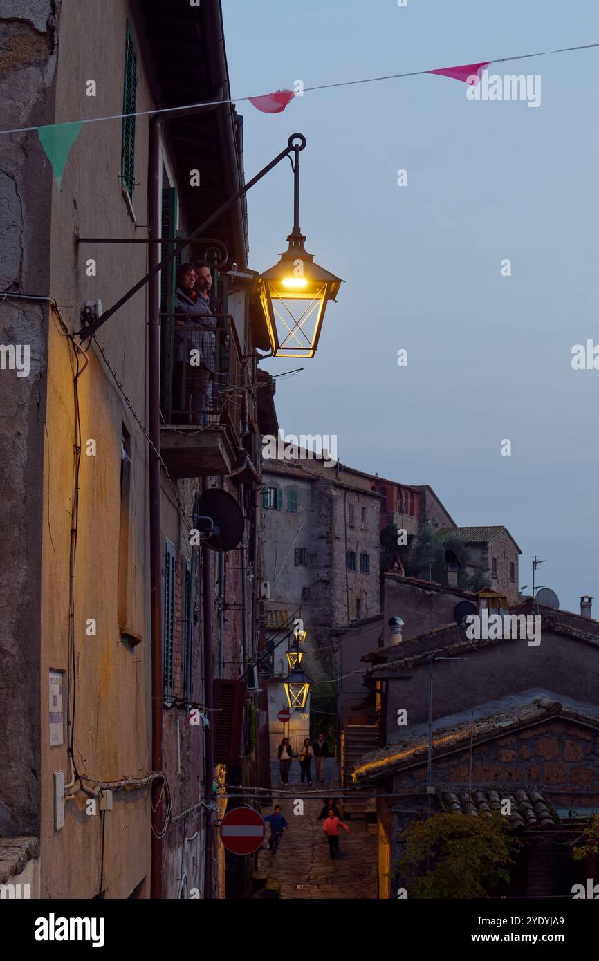 Bunting et lampadaire au crépuscule comme un couple se tiennent sur un balcon avec des gens dans la rue étroite ci-dessous dans la ville de Latera. 27 octobre 2024 Banque D'Images