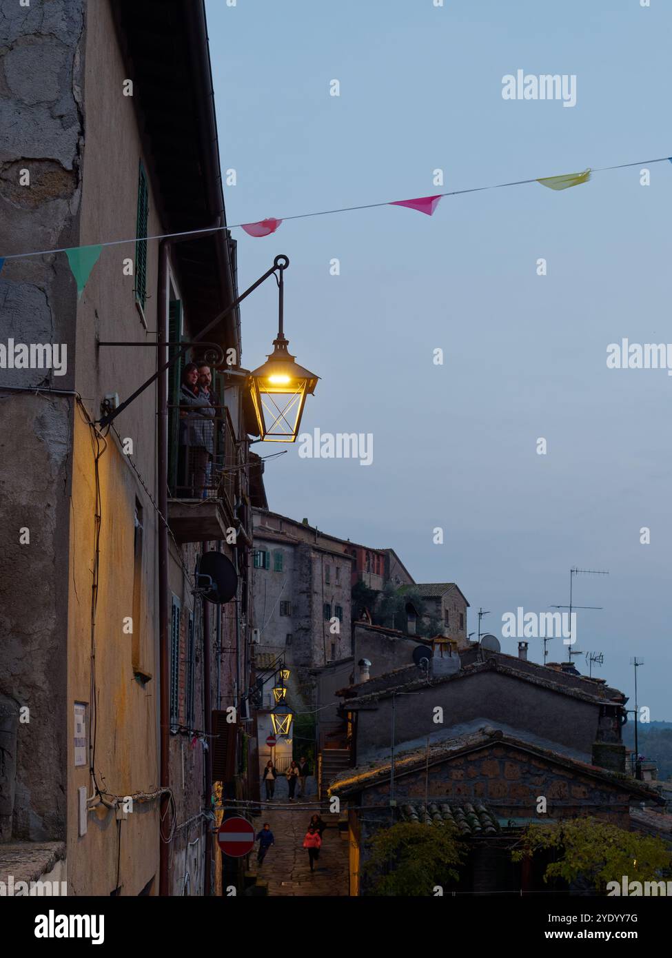 Bunting et lampadaire au crépuscule comme un couple se tiennent sur un balcon avec des gens dans la rue étroite ci-dessous dans la ville de Latera. 27 octobre 2024 Banque D'Images