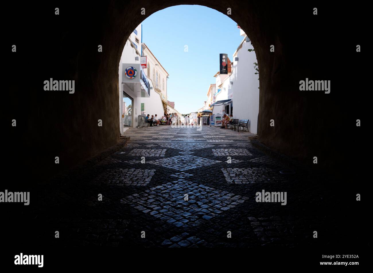 Vue sur le tunnel menant à une rue ensoleillée d'Albufeira, mettant en valeur les pavés à motifs et la vie de la ville animée Banque D'Images