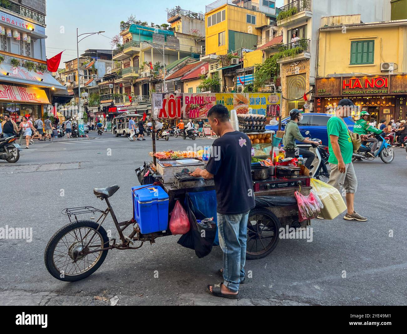 Hanoi, Vietnam : un vietnamien cuisinant de la nourriture sur son chariot à roues au milieu des rues bondées de la ville, des voitures et des motos qui passent à pleine vitesse Banque D'Images