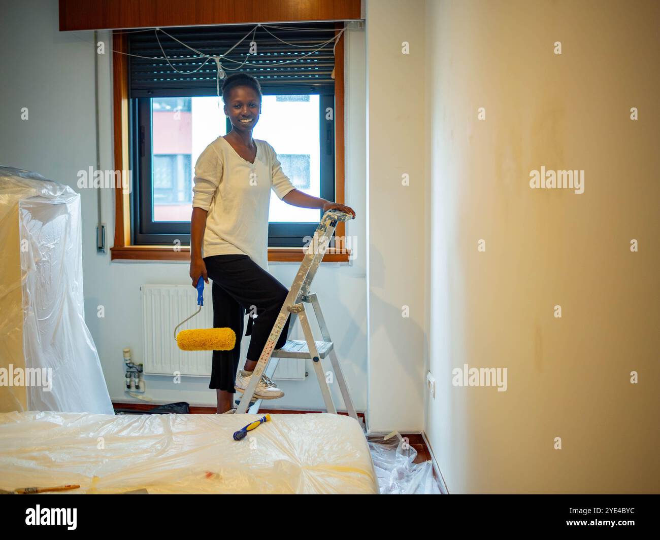 jeune femme sur une échelle avec un rouleau à peinture dans la main souriant regardant la caméra Banque D'Images