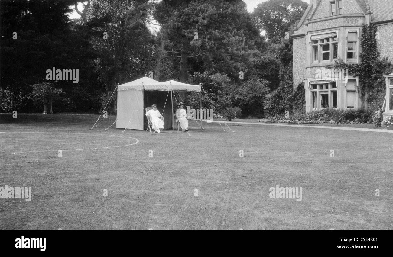 West Sussex, Angleterre. c.1913 – deux femmes membres de la famille Henty, sont assises près d’une petite tente de pavillon sur la pelouse devant Ferring Grange, à Ferring, un village côtier de West Sussex. Ce domaine était la maison d'Edwin Henty, J. P, D.L., F.S.A. (1844-1916), qui avait servi comme haut shérif du Sussex. En 1924, la maison a été convertie en un hôtel à la mode, visité par de nombreuses célébrités dont Edward, le prince de Galles (plus tard connu sous le nom de duc de Windsor). La maison a été détruite par un incendie en octobre 1946. Banque D'Images