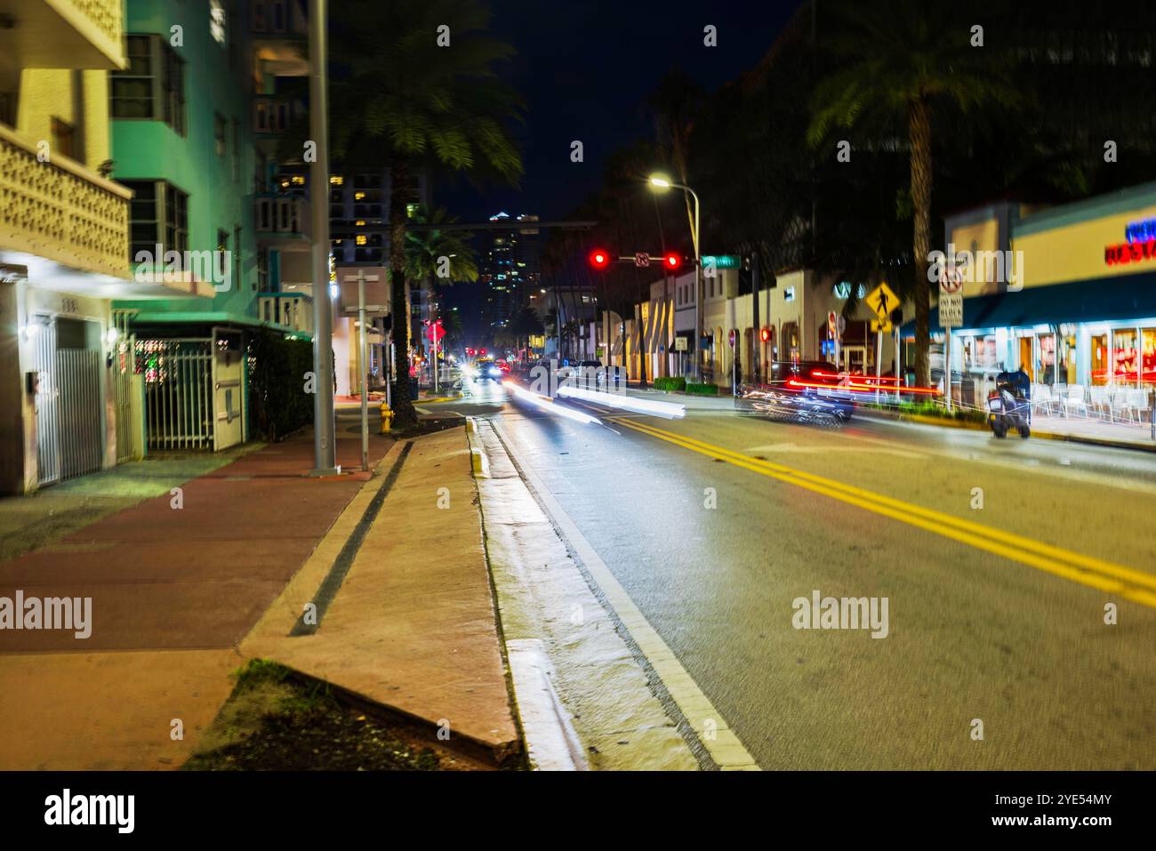 Vue de nuit sur la rue avec des sentiers légers de voitures le long de Collins Avenue à Miami Beach, avec des palmiers flous et des bâtiments en arrière-plan. Banque D'Images