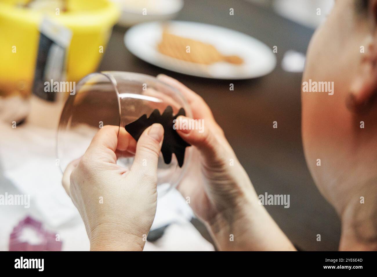 Avec ses mains, une enfant adhère soigneusement une forme d'arbre en feutre noir à un verre tout en étant assis à une table remplie de Cristmas Craft et de friandises. Banque D'Images