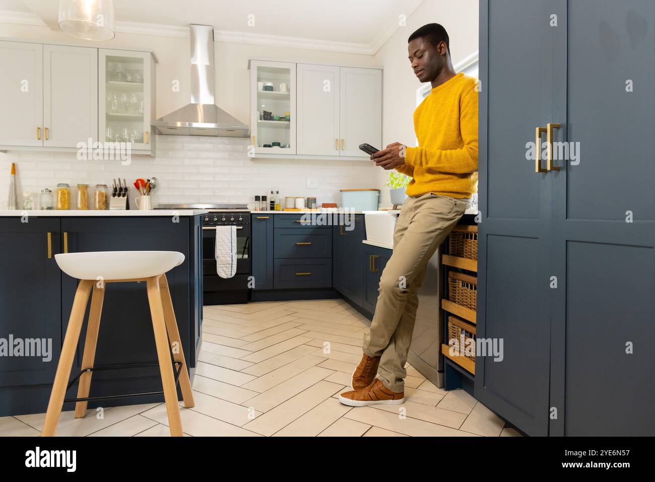 Homme afro-américain dans la cuisine à l'aide d'un smartphone, portant un pull jaune, à la maison Banque D'Images