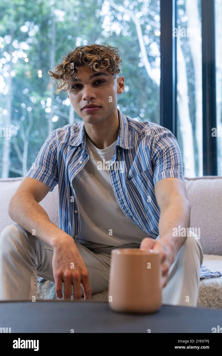 A la maison, jeune homme tenant une tasse à café, se relaxant par une grande fenêtre Banque D'Images