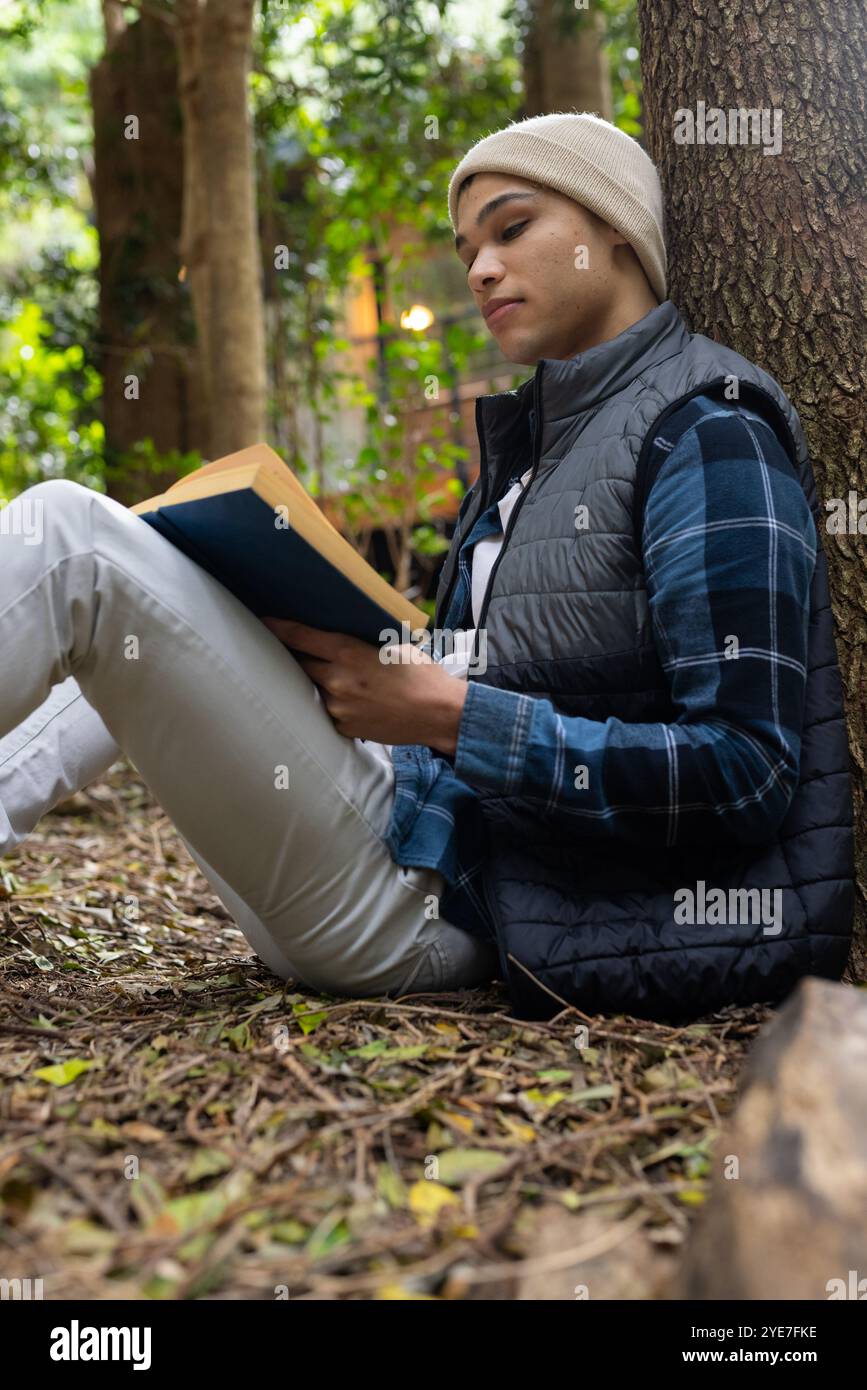 Jeune homme lisant le livre à l'extérieur, se relaxant contre l'arbre dans la forêt paisible Banque D'Images