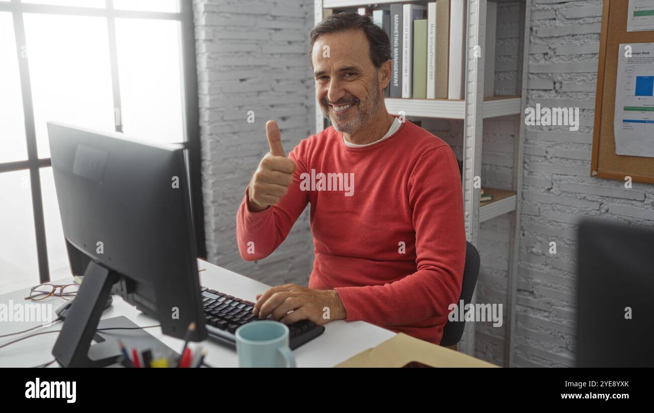 Homme hispanique souriant au bureau dans un bureau moderne montrant le pouce vers le haut geste au milieu des livres et des documents dans un intérieur lumineux Banque D'Images