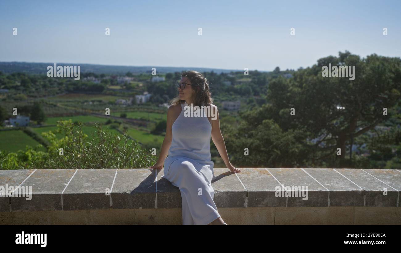 Jeune femme hispanique dans une tenue élégante profitant de la vue panoramique depuis un rebord de pierre à locorotondo, pouilles, italie, entourée de verdure luxuriante et dist Banque D'Images