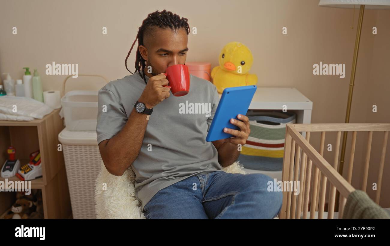 Jeune homme avec des tresses est assis dans la chambre à boire de la tasse tout en lisant la tablette à côté du lit de bébé dans l'environnement de la maison confortable Banque D'Images