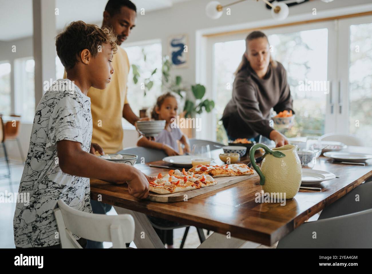 Garçon gardant le tableau de pizza pendant que les parents dressent la table à manger pour le déjeuner à la maison Banque D'Images