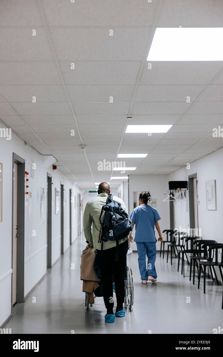 Vue arrière d'un homme mûr marchant avec sa mère assise sur un fauteuil roulant dans le couloir de l'hôpital Banque D'Images