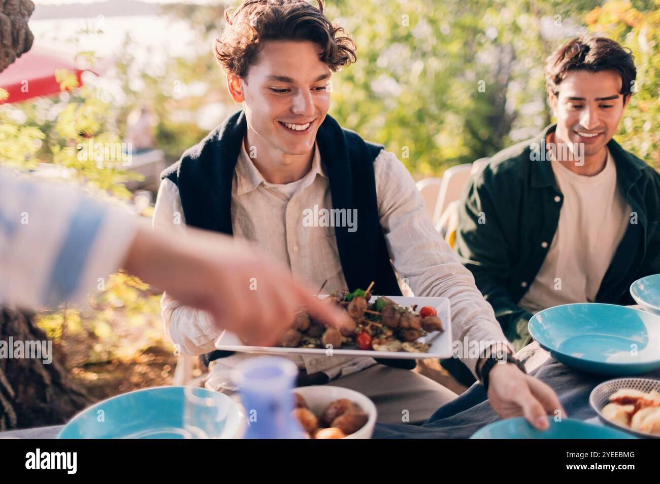 Homme souriant tenant le plateau d'apéritif tout en étant assis avec un ami mâle à la table à manger pendant la fête Banque D'Images