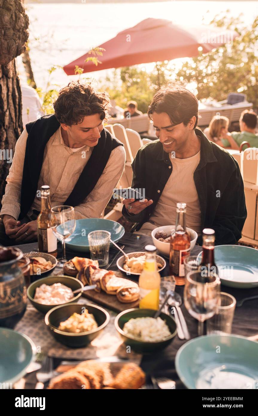 Jeune homme souriant partageant un téléphone intelligent avec un ami masculin assis à la table à manger pendant la soirée du dîner Banque D'Images
