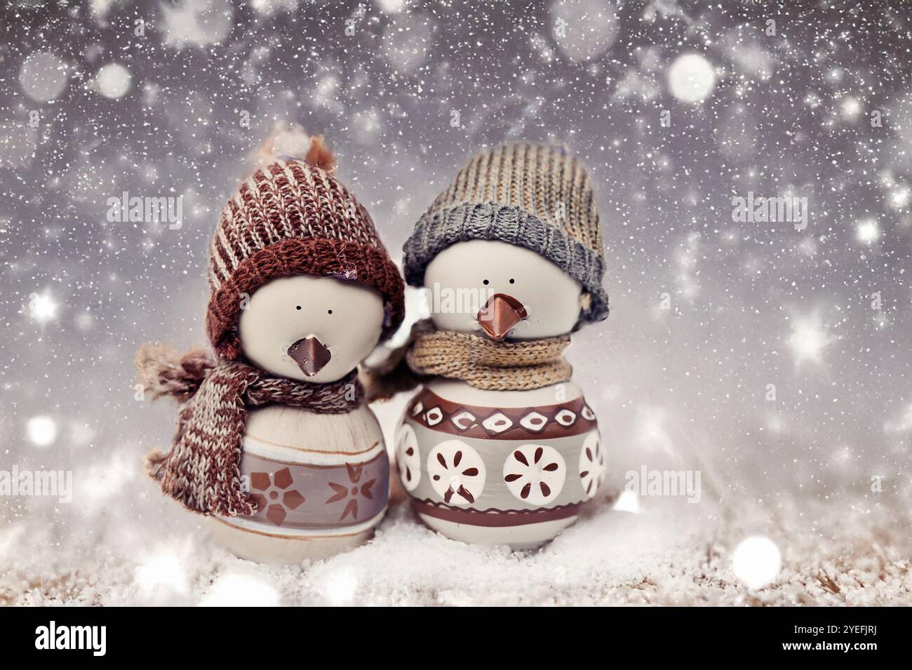 Deux adorables figurines d'oiseau de neige habillées de chapeaux en tricot douillets et de foulards, debout côte à côte dans un cadre enneigé avec des lumières bokeh étincelantes, Banque D'Images