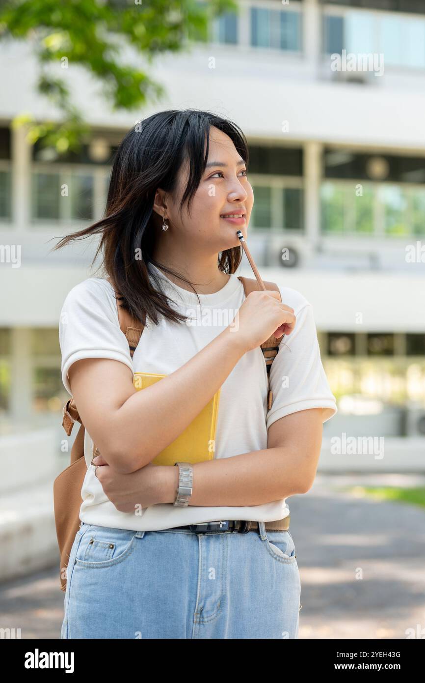 Une étudiante d'université féminine asiatique jolie et réfléchie dans des vêtements décontractés avec un sac à dos se tient sur son campus universitaire, tenant un crayon à son menton comme s. Banque D'Images