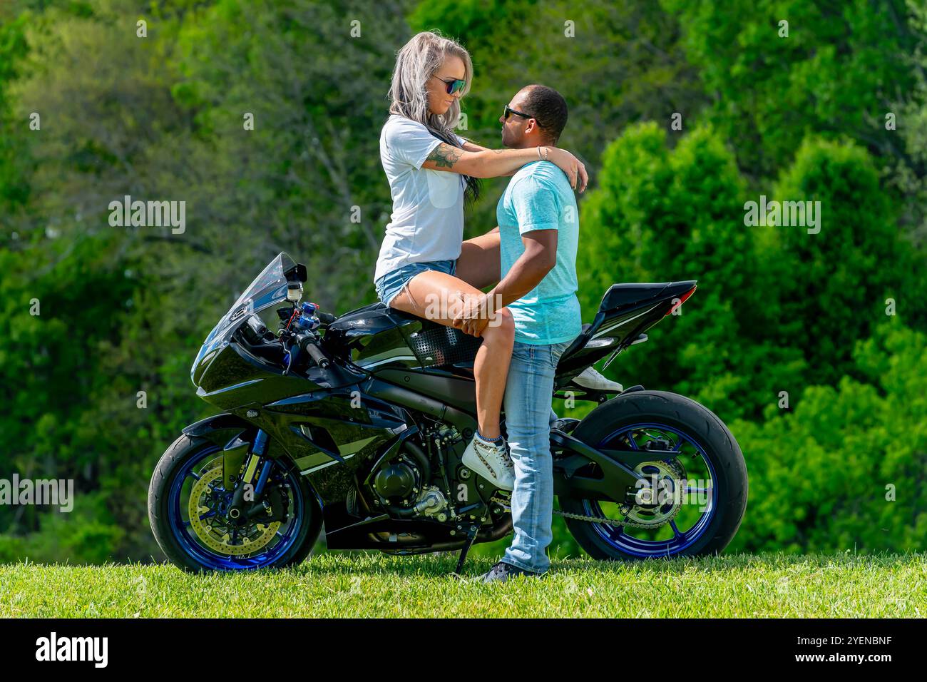 Un couple multiracial pose avec amour à côté de leur moto dans un champ verdoyant et luxuriant. Embrassés, ils sourient avec fierté, capturant à la fois leur lien profond A. Banque D'Images