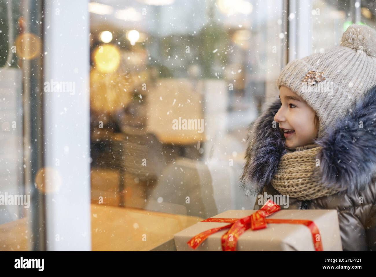 Portrait de fille joyeuse avec une boîte cadeau pour Noël près de la fenêtre de la boutique de verre en hiver avec la neige sur le marché de fête avec des décorations et des lumières. WA Banque D'Images