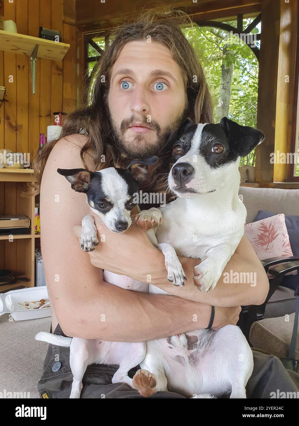 Jeune homme avec de grands yeux bleus et de longs cheveux blonds tenant fermement à sa poitrine deux petits chiens noirs et bruns, un petit chihuahua et un terrier Banque D'Images