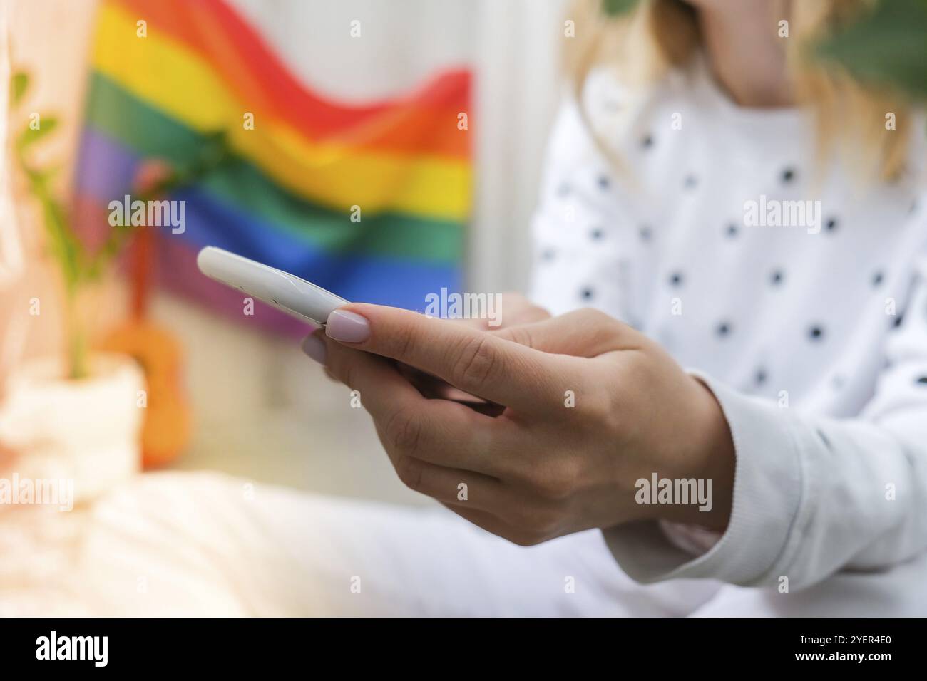 Jeune femme hippie millénaire assise sur un balcon avec téléphone mobile. Drapeau arc-en-ciel LGBTQ sur fond. Envoi de SMS par téléphone, appel vidéo, prise de selfie. Blogg Banque D'Images