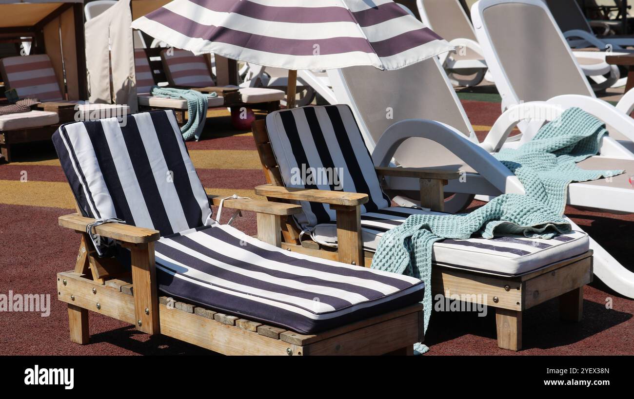 deux chaises longues en bois pour enfants avec couverture en coton doux rayé à côté des chaises longues pour adultes sous parasol en tissu, piscine vide pour la détente Banque D'Images