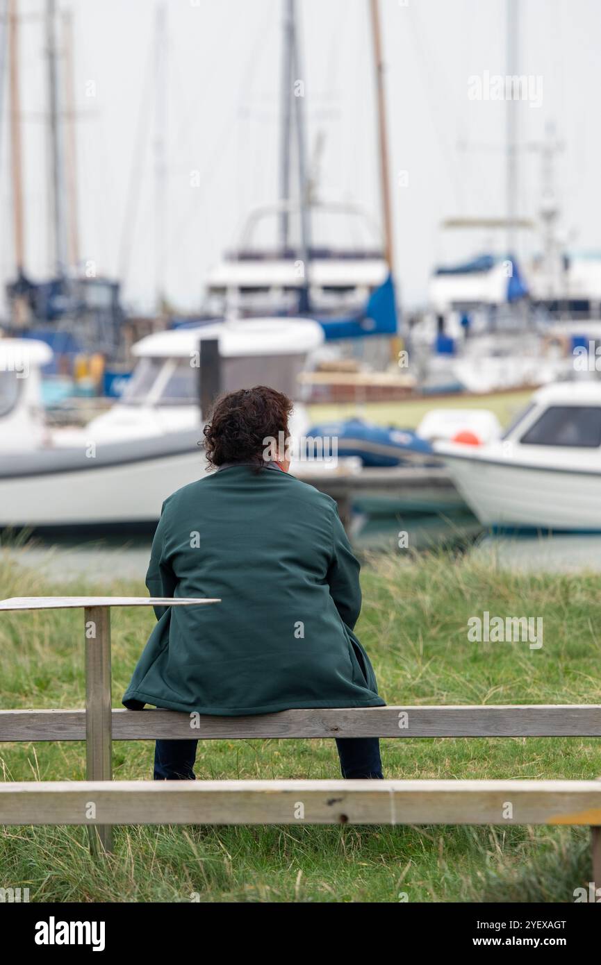 Dame âgée ou d'âge moyen assise seule sur un banc en bois au bord de la mer donnant sur un port de plaisance animé à Yarmouth sur l'île de wight, Royaume-Uni Banque D'Images