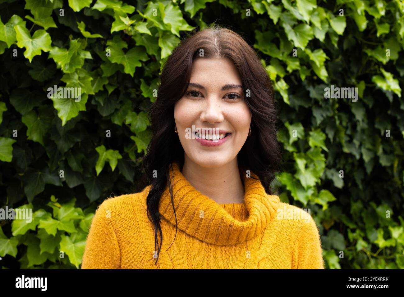 Femme souriante en pull jaune debout à l'extérieur contre des feuilles vertes luxuriantes Banque D'Images