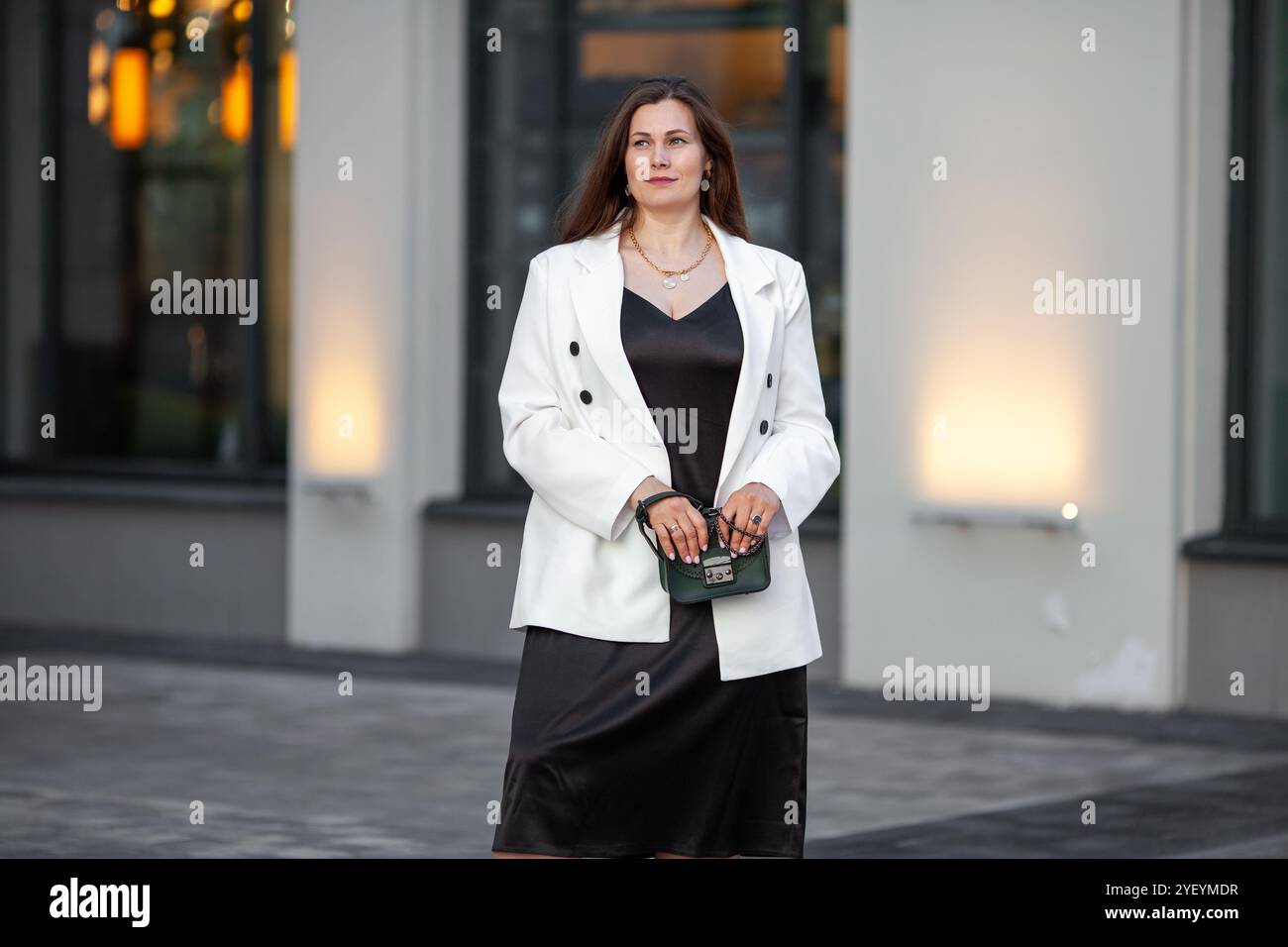Grande taille femme élégante se tient à l'extérieur dans un cadre urbain à la ville, portant une robe noire à la mode avec une tenue blazer double croisée blanche. Modèle avec Banque D'Images