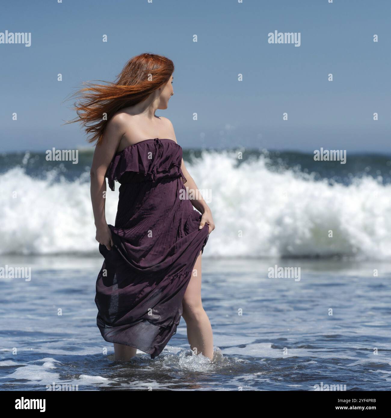 Redhead femme en robe longue avec genou haut ourlet debout cheville profonde dans l'eau sur la plage et la tête tournée, regardant par-dessus l'épaule à la mer en prévision de Banque D'Images