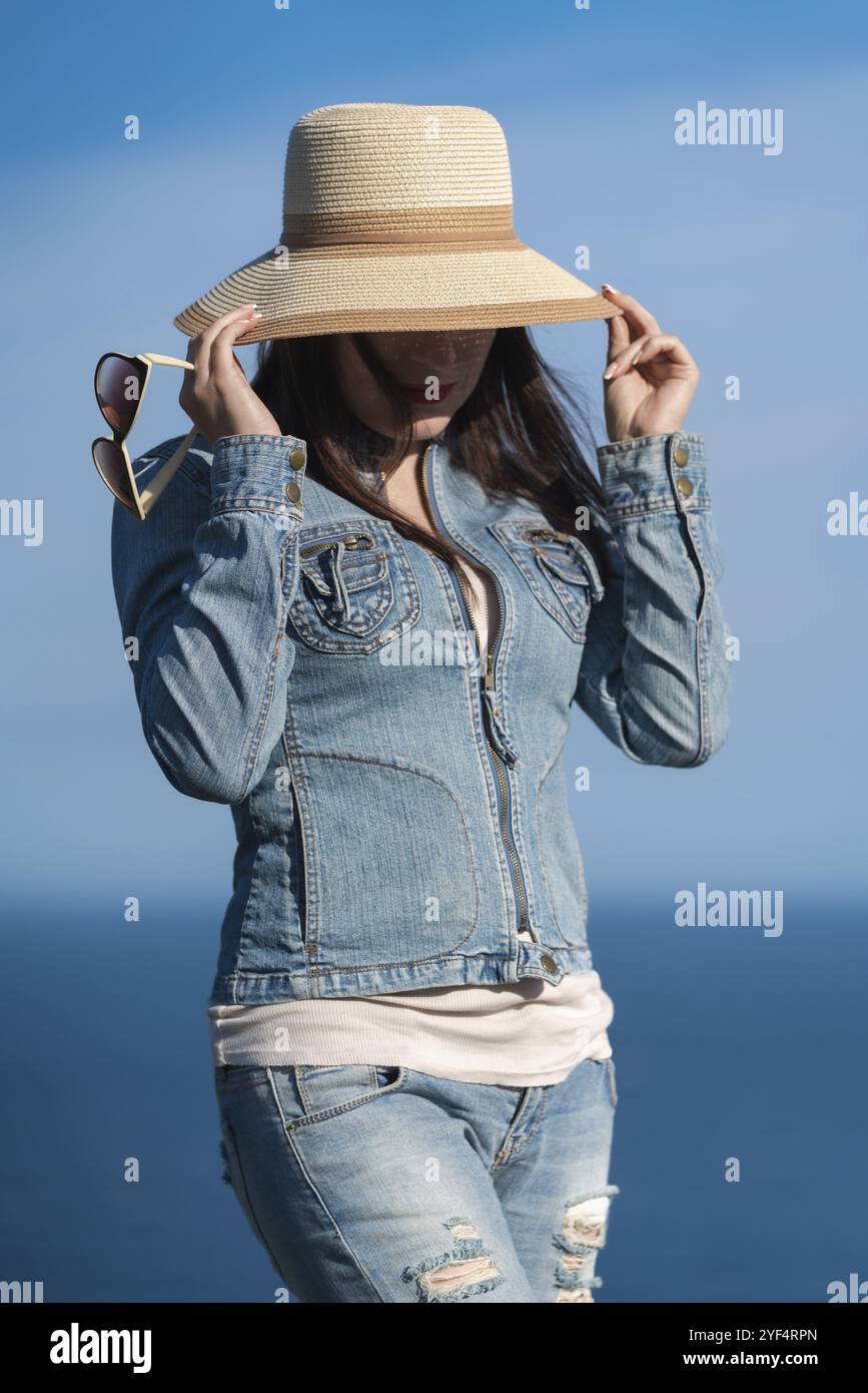 Portrait de femme hipster en veste denim et jeans se cachant avec chapeau de paille sur son visage. Femelle adulte posant sur fond de ciel bleu sans nuages et o Banque D'Images