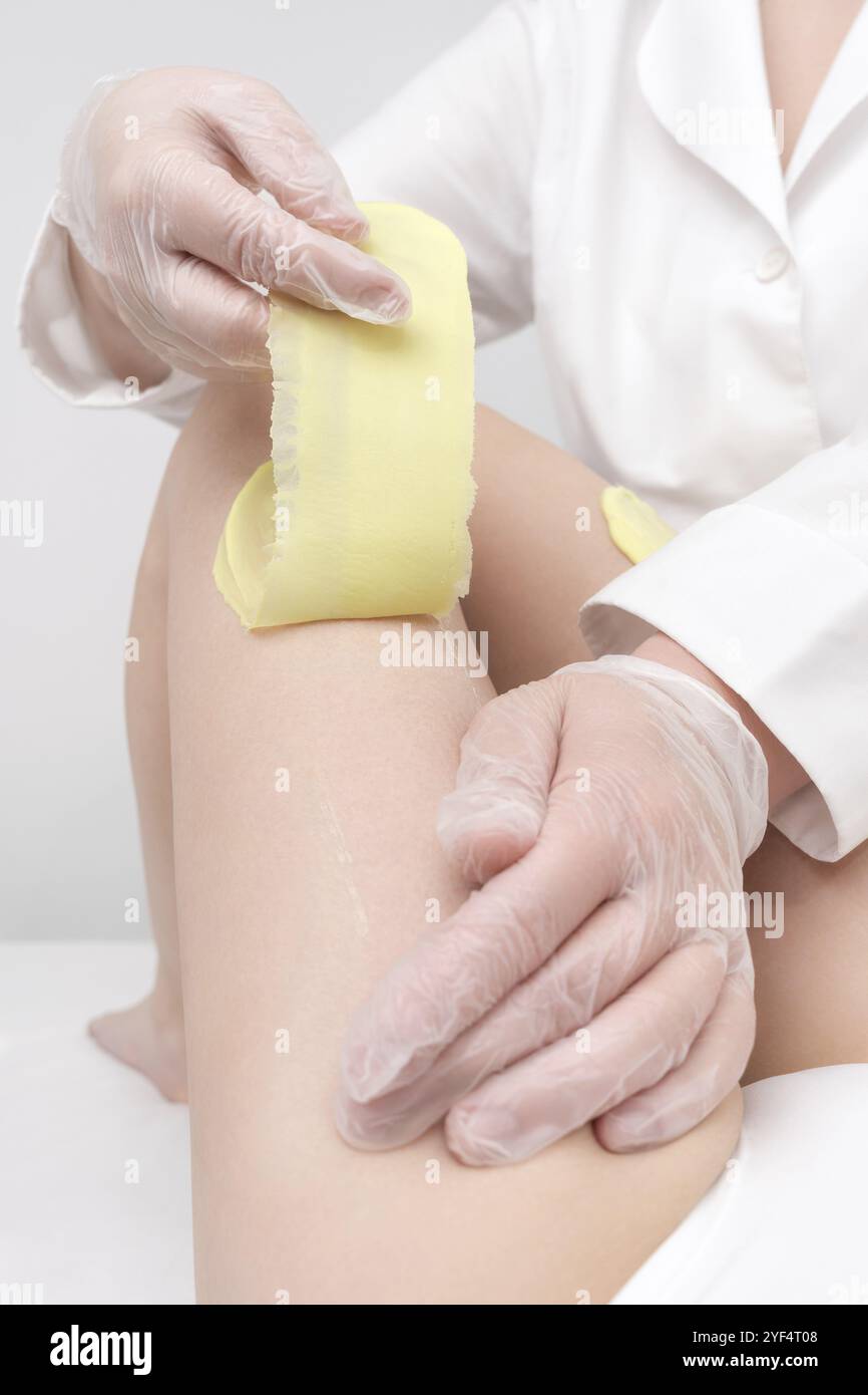 Procédure d'épilation, cosmétologue mains dans des gants enlevant les poils sur la jambe des femmes. Processus d'épilation avec de la cire chaude verte dans un salon de beauté professionnel. Partie o Banque D'Images