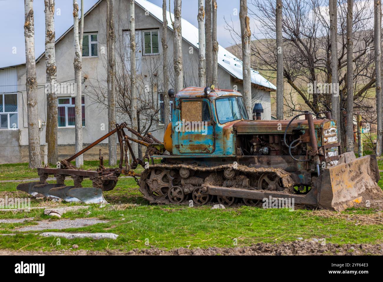 Bulldozer agricole rustique avec une charrue contre une maison de deux étages et une rangée d'arbres Banque D'Images