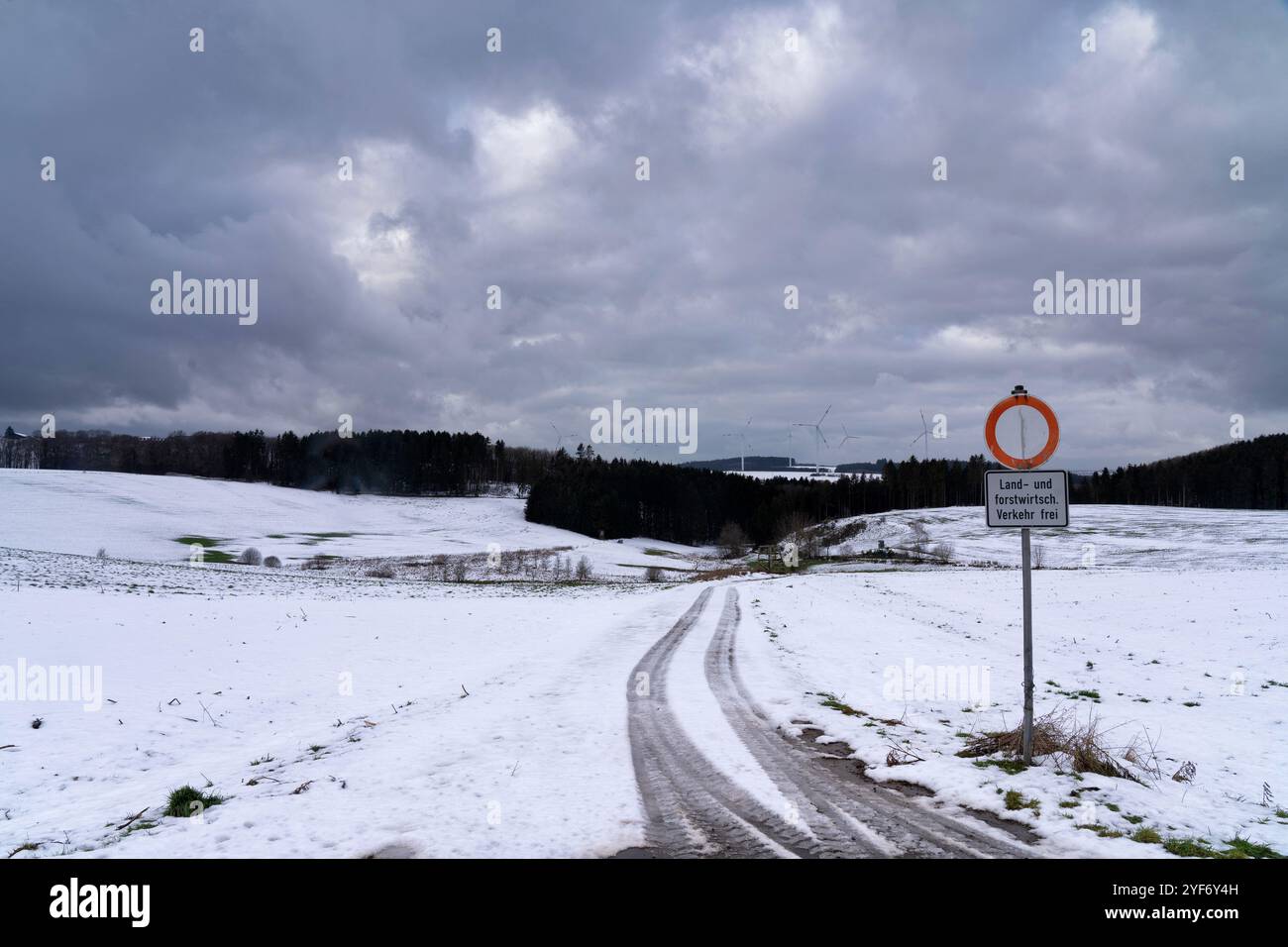 Un paysage hivernal présente une route enneigée serpentant vers une forêt lointaine. Le ciel est rempli de nuages sombres, faisant allusion au changement de temps. Banque D'Images