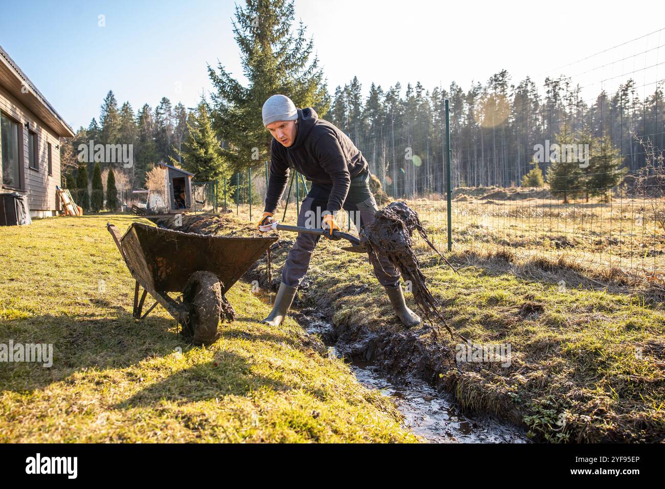 Homme en équipement de protection travaillant inlassablement pour draguer une tranchée boueuse avec une pelle et une brouette dans une arrière-cour au début du printemps. Banque D'Images