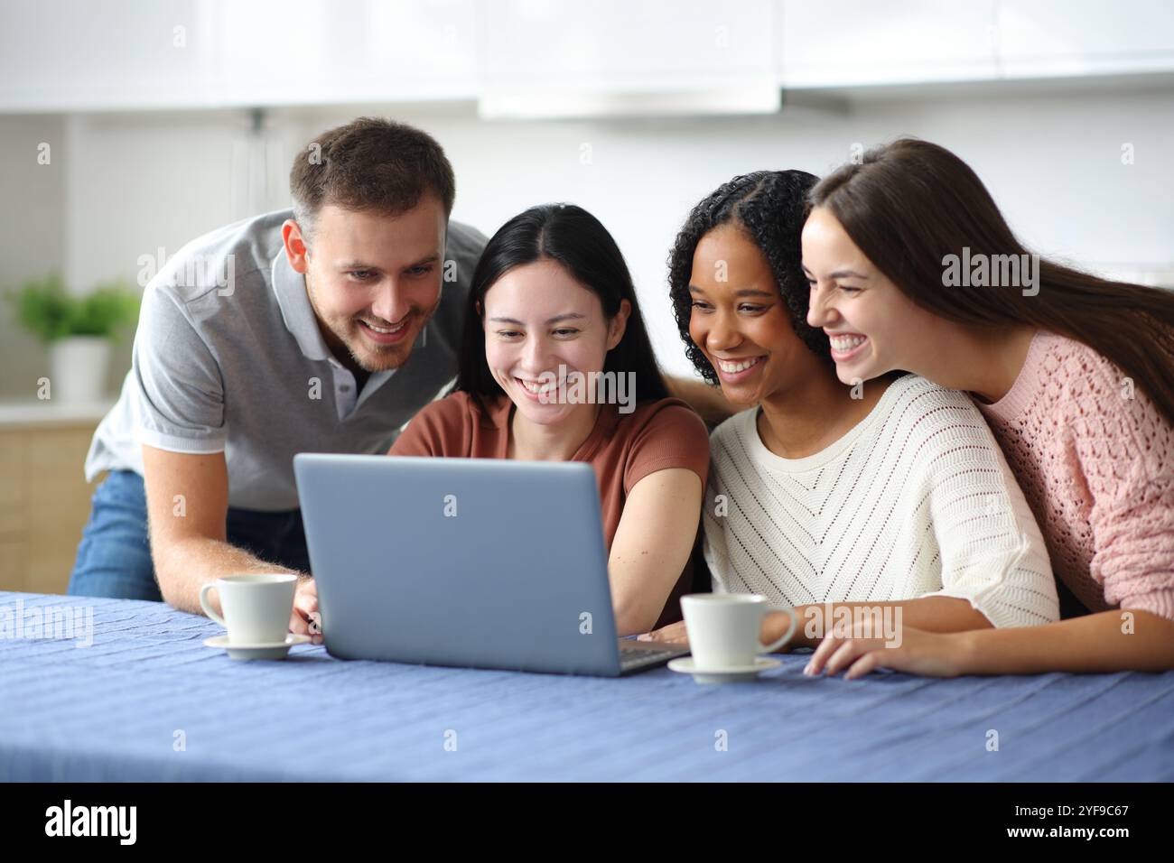 Quatre amis heureux interraciaux vérifiant les médias en ligne d'ordinateur portable dans la cuisine à la maison Banque D'Images