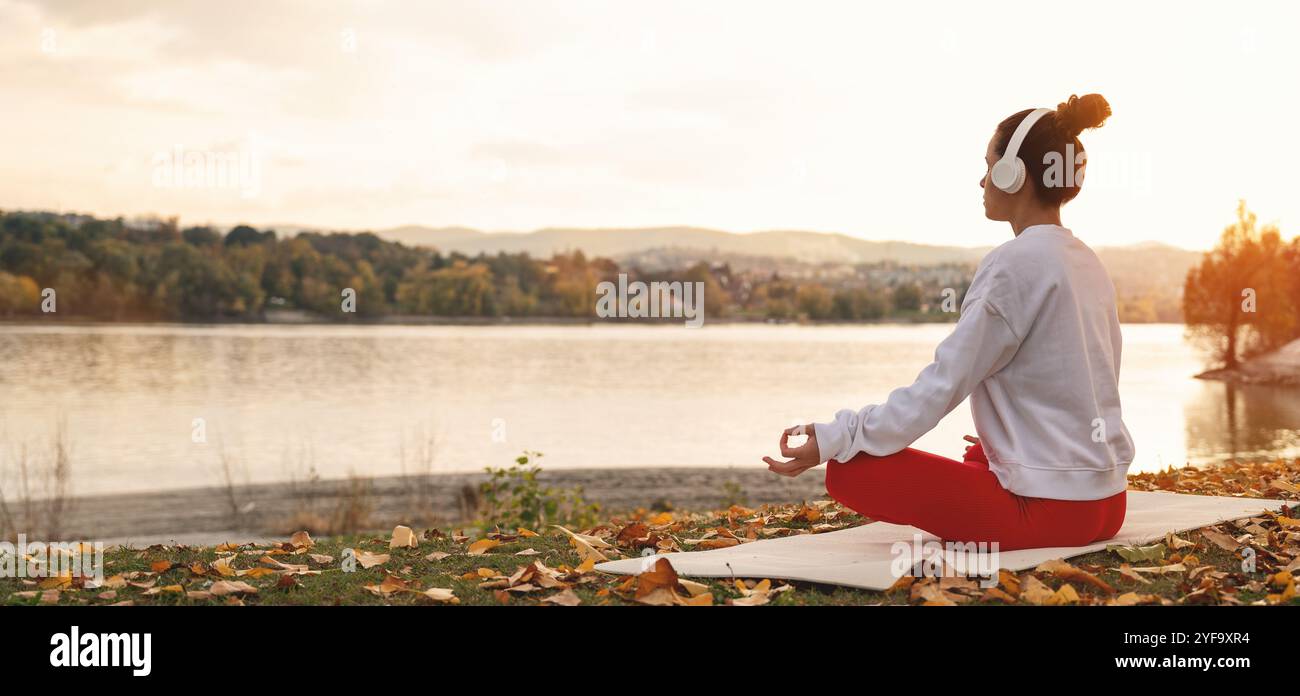 La femme dans les écouteurs est assise dans la pose de lotus devant la rivière d'automne dans la nature et méditer pendant le coucher du soleil. Mode de vie sain et évasion urbaine. Banque D'Images