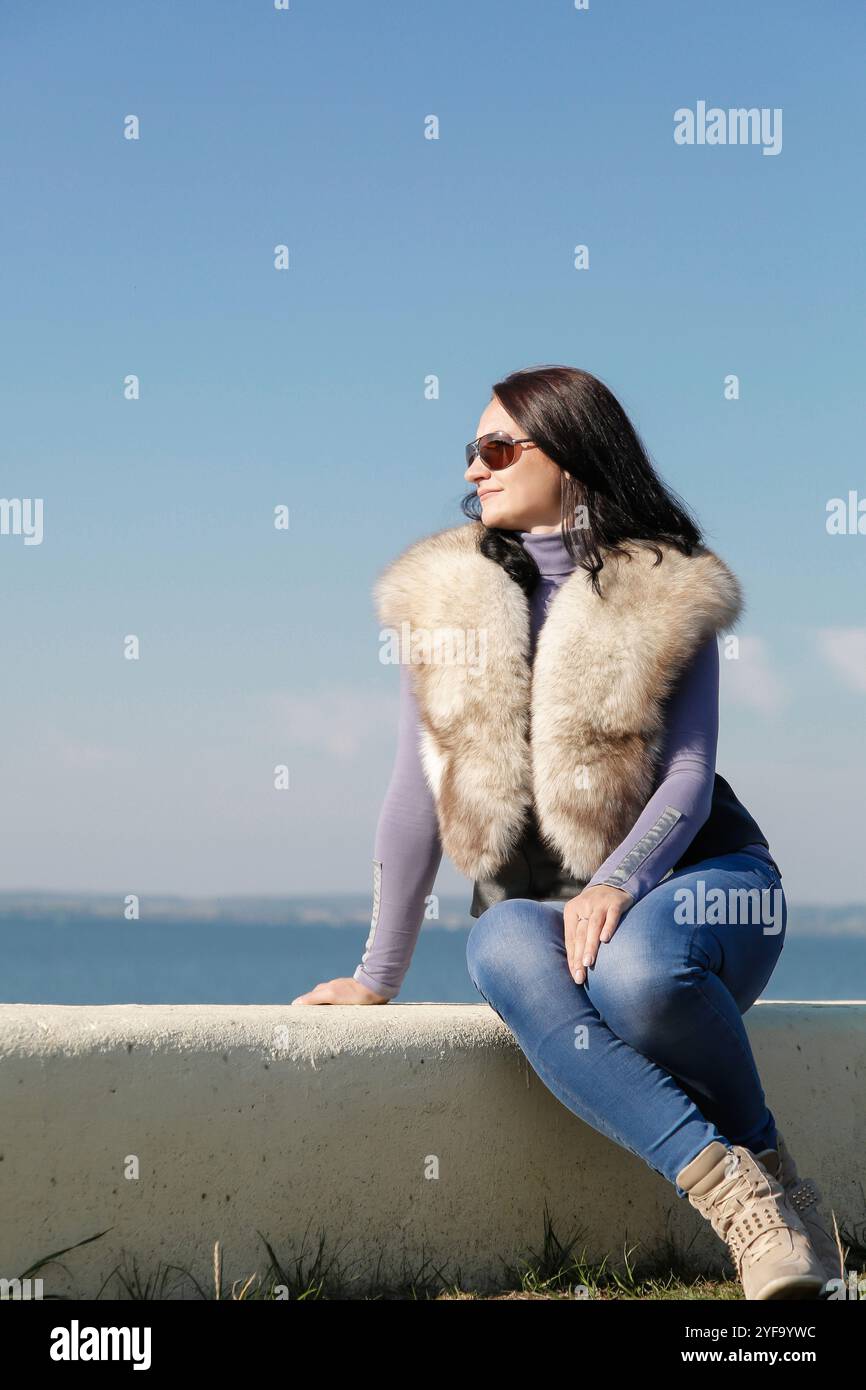 Une femme élégante et à la mode dans un gilet de fourrure se trouve sur une jetée sur le fond de la mer Banque D'Images