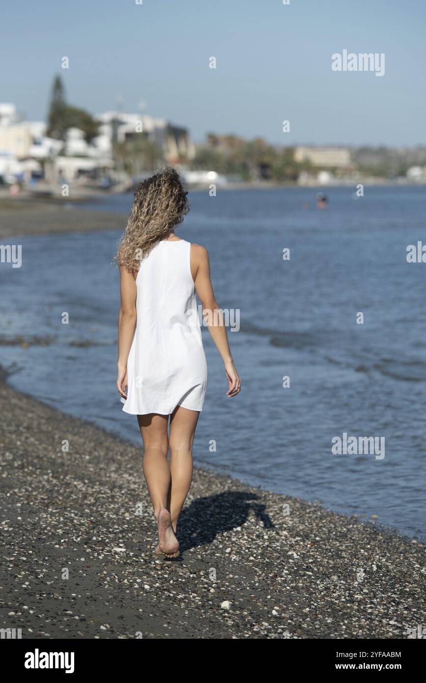 Jeune femme méconnue portant une robe d'été blanche marchant sur la plage. Style de vie en extérieur. Marche dans le littoral Banque D'Images