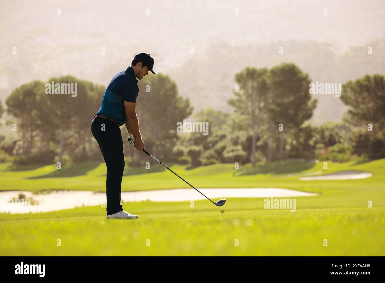 Un golfeur professionnel se concentre sur un tee shot avec un pilote, près d'un bunker de sable, incarnant la concentration et le défi sur le terrain de golf. Banque D'Images