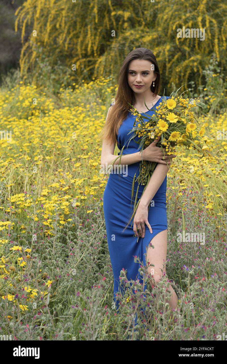 Belle jeune femme caucasienne attrayante, vêtue de vêtements bleus dans les champs couverts de fleurs printanières jaunes. Concept d'accueil du printemps Banque D'Images