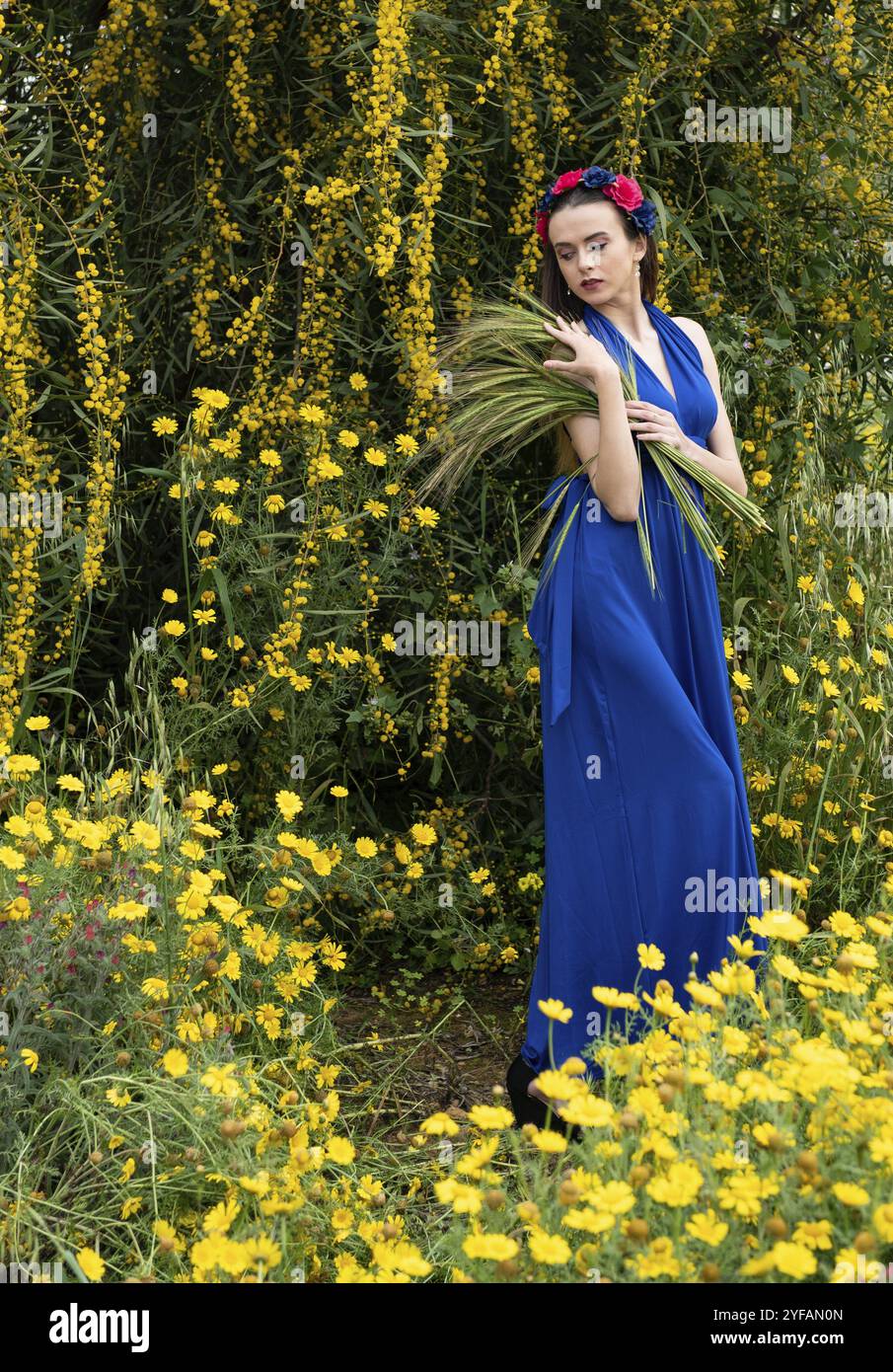 Young caucasian belle dame, habillée de vêtements bleu et avec une couronne de fleurs sur la tête dans les champs couverts de printemps jaune bleu ... Banque D'Images
