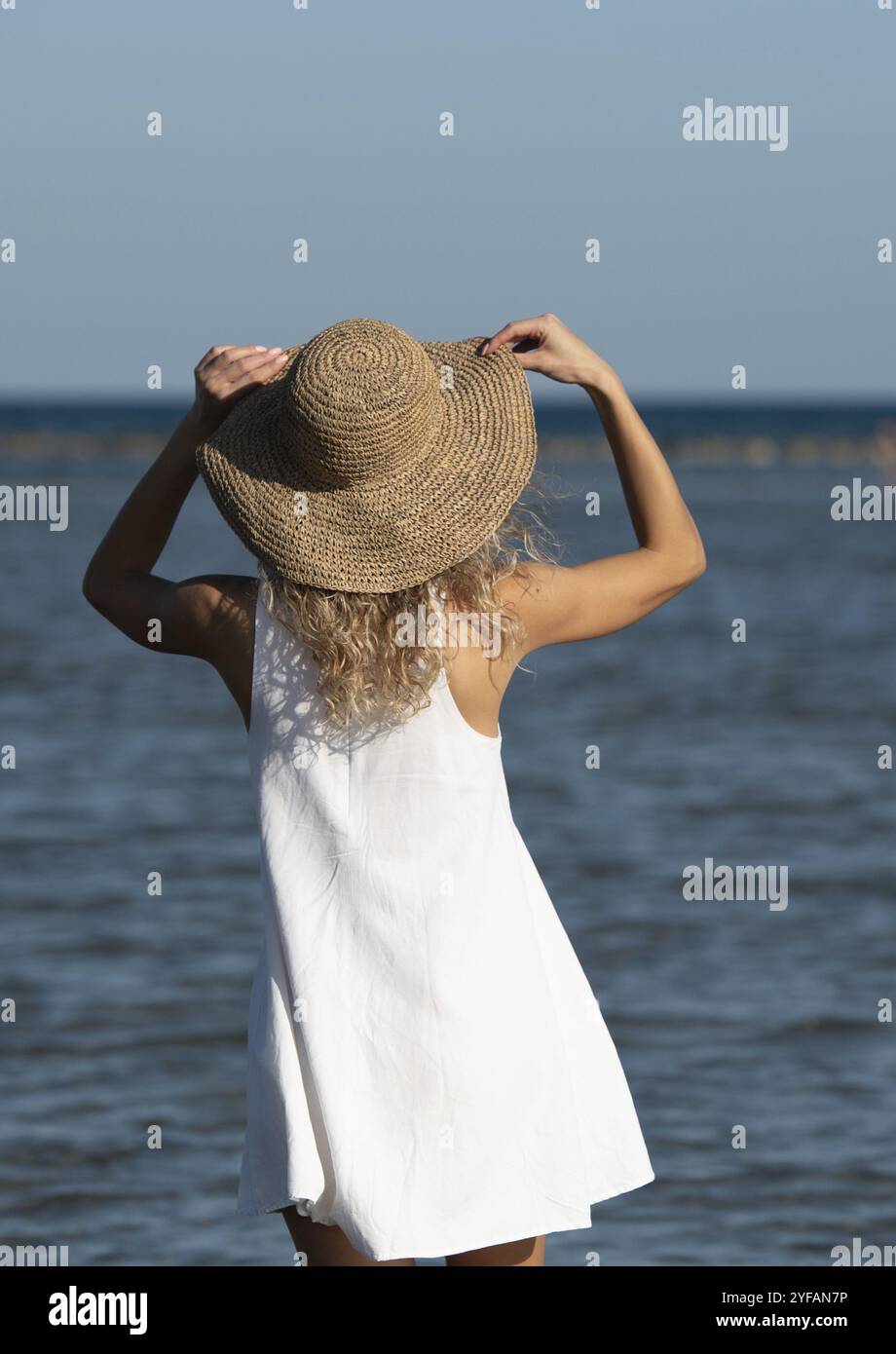 Jeune femme méconnue portant une robe d'été blanche et un chapeau marchant dans la plage. Style de vie en extérieur. Marche dans le littoral Banque D'Images
