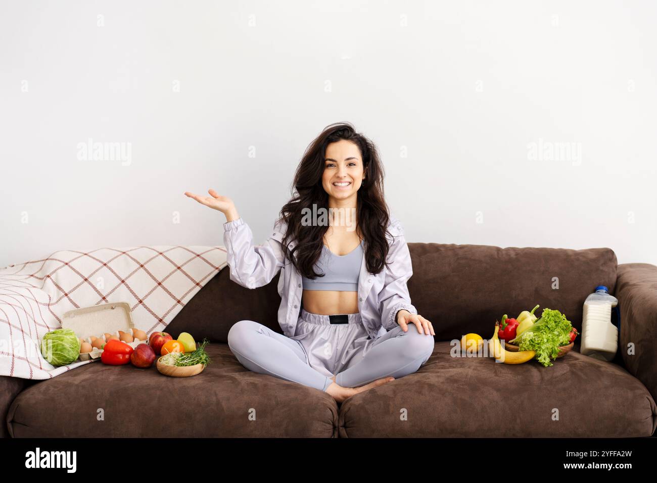 Jeune femme souriante assise sur son canapé avec les jambes croisées, faisant des gestes vers une diffusion d'aliments et de boissons sains Banque D'Images